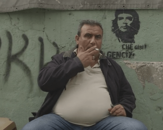Screen shot a man sits in front of PKK grafiti: Note who the picture is next to this man: Che Guevera